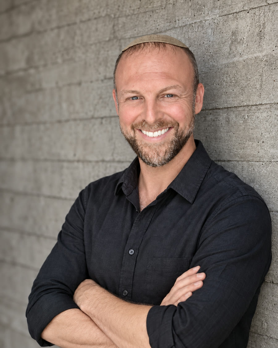 Yakir Hyman smiling, wearing a dark button-down shirt and kippah, leaning against a light stone wall